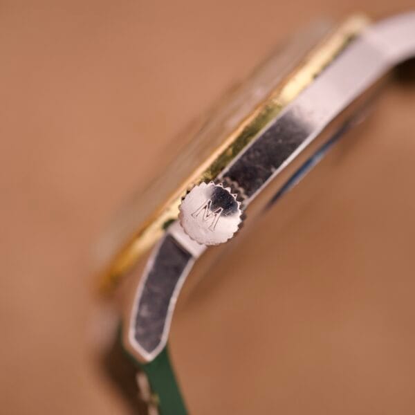 Close-up of a silver watch crown engraved with an M, attached to a gold and silver Wyler Dynawind Heavy Duty 660 Life Guard case.