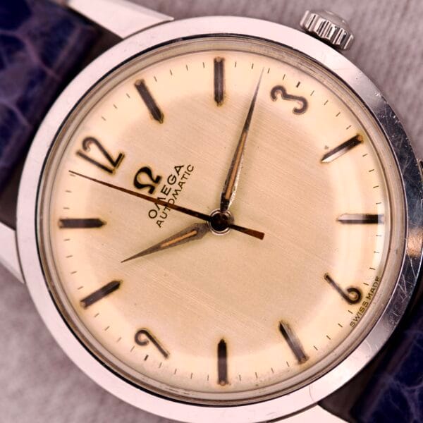 A 1959 Omega Ref. 14773-1 SC Linen Dial C. 470 wristwatch featuring a cream linen dial, black markers, and a blue leather strap.