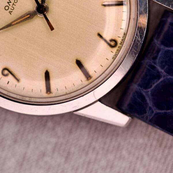 Close-up of a 1959 Omega Ref. 14773-1 SC Linen Dial C. 470 vintage wristwatch featuring a cream linen dial, black markers, and a blue textured leather strap.