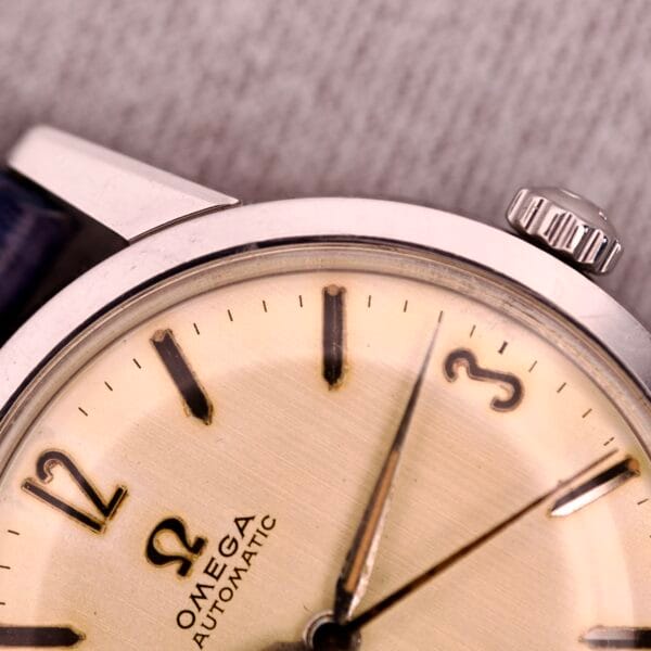Close-up of a 1959 Omega Ref. 14773-1 SC automatic wristwatch with Caliber 470, showcasing its linen dial, intricate hands, and crown on a textured surface.
