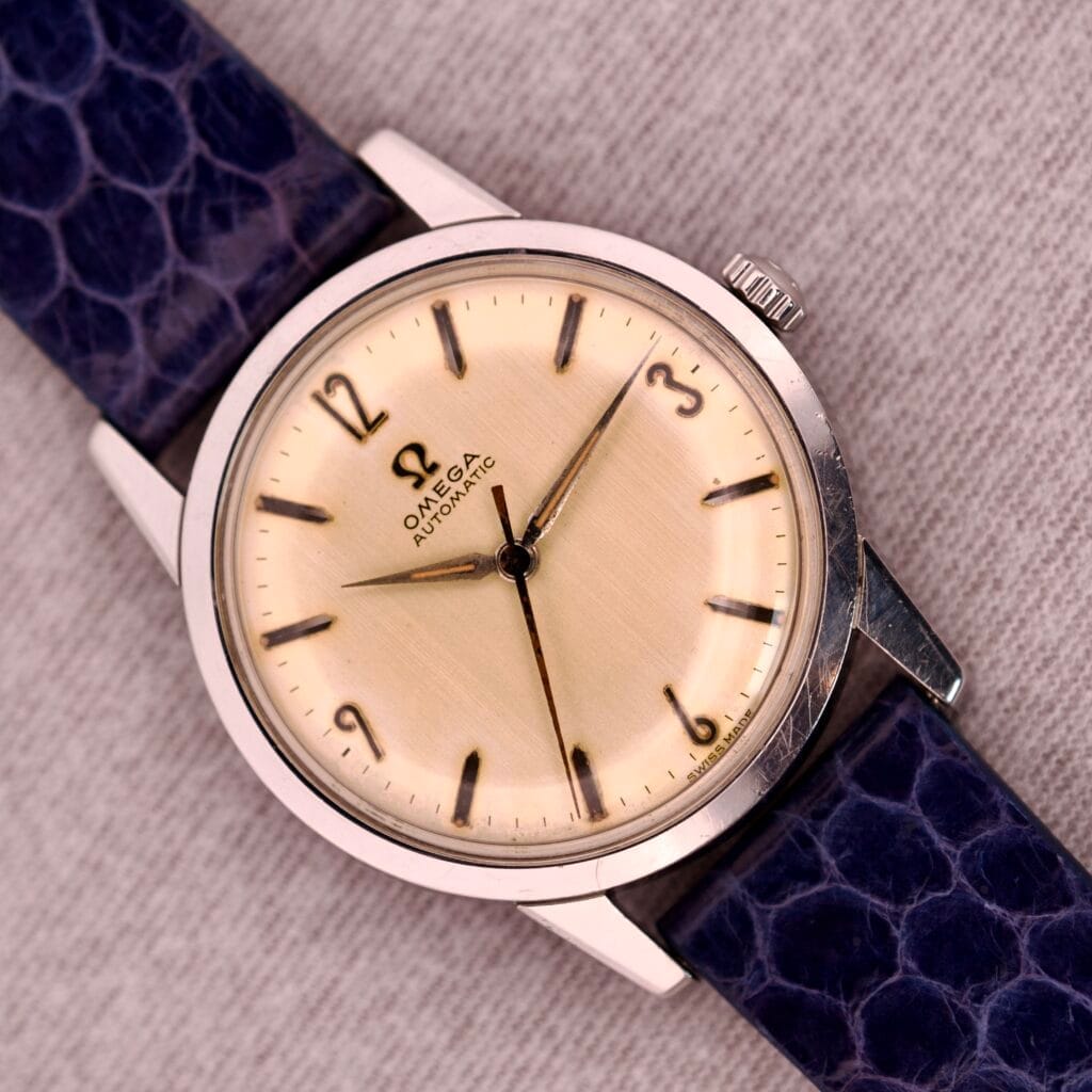 A 1959 Omega Ref. 14773-1 SC Linen Dial C. 470 wristwatch with a cream linen dial and blue textured strap, showcased on a light fabric background.