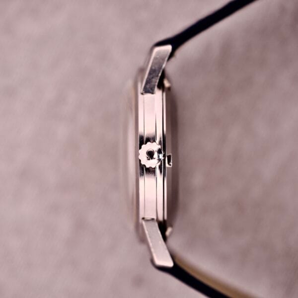 Side view of a 1959 Omega Ref. 14773-1 SC Linen Dial C. 470 wristwatch with a black strap, shown on a textured light background.