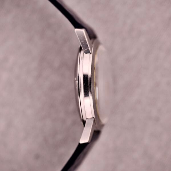 Side view of a 1959 Omega Ref. 14773-1 SC Linen Dial C. 470 silver wristwatch with black strap, shown on a light textured background.