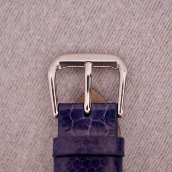 Close-up of a silver buckle on a textured purple leather strap atop beige fabric, echoing the refined detailing of the 1959 Omega Ref. 14773-1 SC Linen Dial C. 470 and its elegant linen dial design.