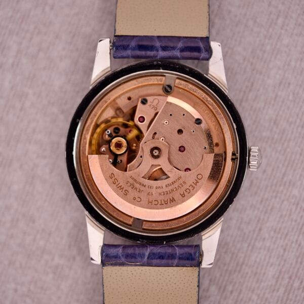 Close-up of a 1959 Omega Ref. 14773-1 SC Linen Dial C. 470 wristwatch showing its exposed Caliber 470 mechanical movement and blue strap, displayed on a light linen dial fabric background.