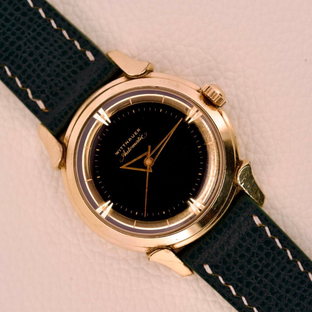 A Wittnauer Automatic Black Gilt Dial Cal. 11ARG wristwatch with a dark leather strap is displayed on a white textured background.