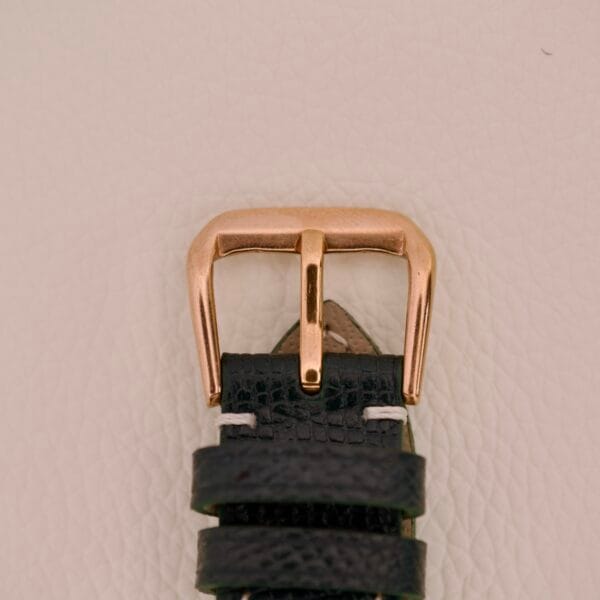 A gold buckle on a black leather strap, paired with the Wittnauer Automatic Black Gilt Dial Cal. 11ARG, rests on a textured white background.