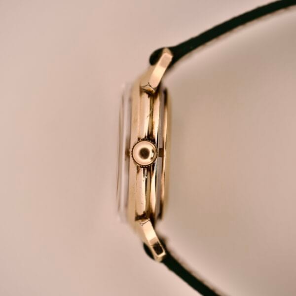 Side view of a Wittnauer Automatic Black Gilt Dial Cal. 11ARG gold wristwatch with black strap, highlighting its crown, slim profile, and elegant black gilt dial against a light background.