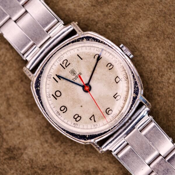 A Tudor Cushion Case Trench Watch Circa 1930s with a silver band and aged white dial rests on a brown textured surface.