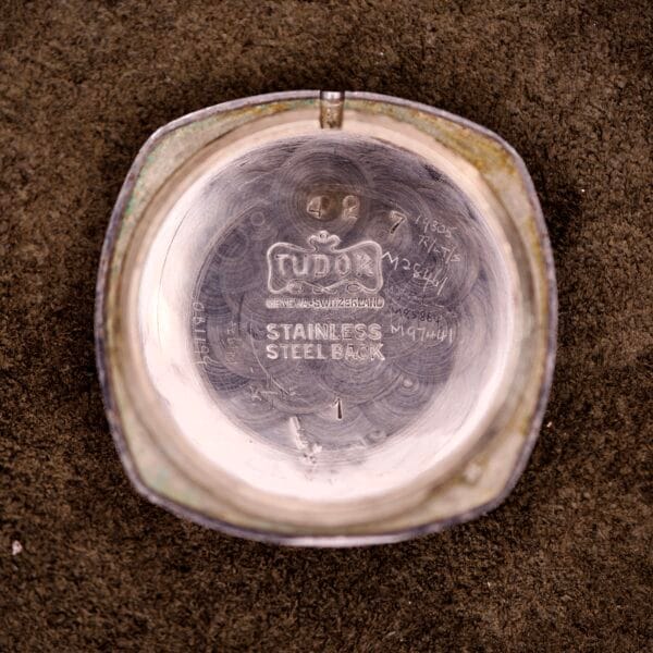 Inside the back of a Tudor Cushion Case Trench Watch Circa 1930s, engraved logos, text, and handwritten markings are visible.