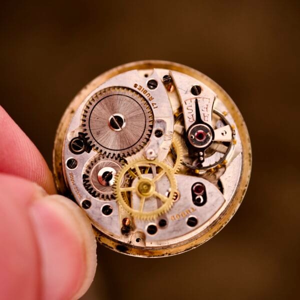 A close-up of fingers holding the exposed Tudor Cushion Case Trench Watch Circa 1930s mechanism, with visible gears and springs reflecting classic craftsmanship.