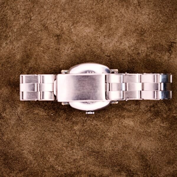 The Tudor Cushion Case Trench Watch circa 1930s is a silver metal wristwatch with a cushion case and closed clasp, displayed on a brown suede surface.