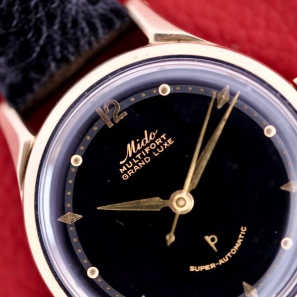Close-up of a Mido Multifort Grand Luxe Super Automatic 14k Gold Capped wristwatch with black dial, gold hands, and Grand Luxe text, highlighting its vintage design and precision.
