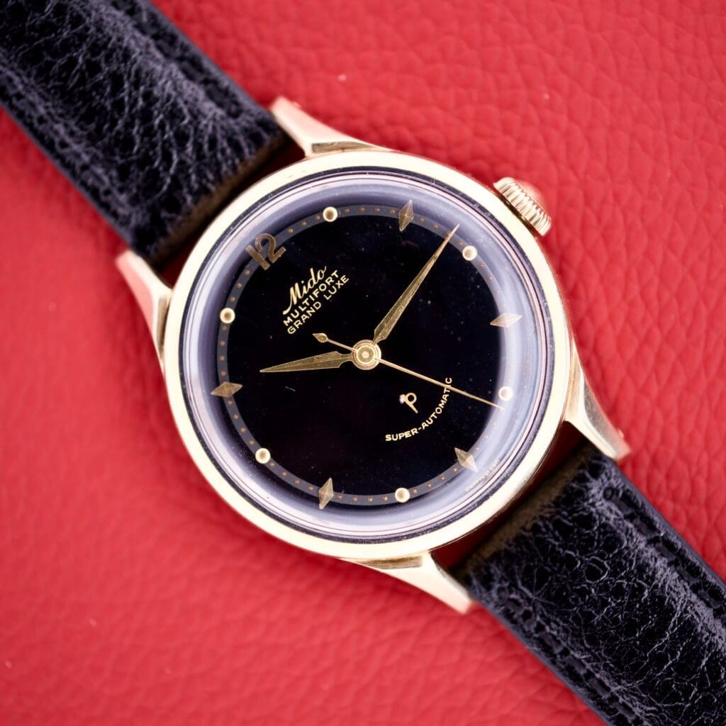 The Mido Multifort Grand Luxe Super Automatic 14k Gold Capped watch is elegantly displayed with a black face and leather strap on a red textured background.