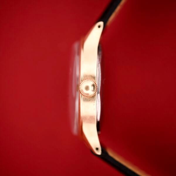 Side view of the Mido Multifort Grand Luxe Super Automatic 14k Gold Capped wristwatch against a red background, highlighting its crown, lugs, and black strap—an elegant display of luxury.