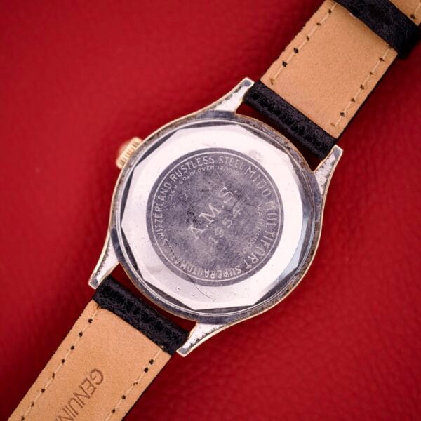 Back of a Mido Multifort Grand Luxe Super Automatic 14k Gold Capped watch with leather straps, showing engraved text on a silver case against a red background.
