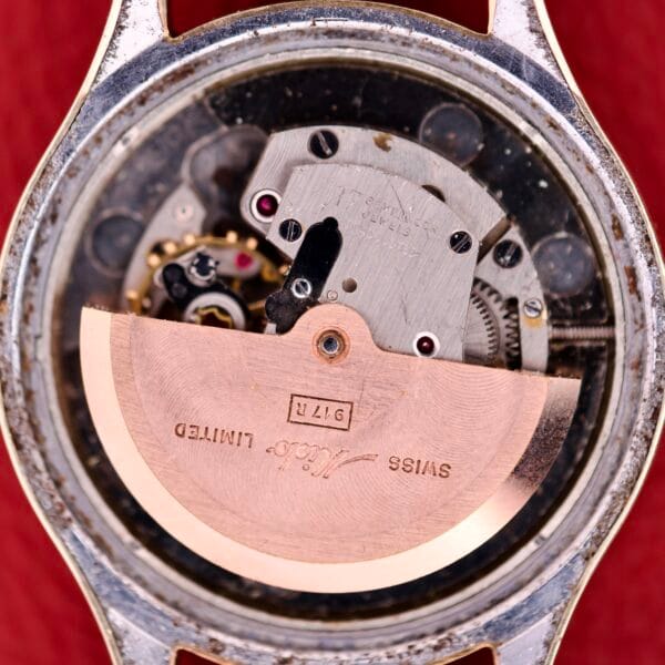 Close-up of the Mido Multifort Grand Luxe Super Automatic 14k Gold Capped watch movement with visible gears and engraved rotor on a red background, highlighting its Swiss craftsmanship.