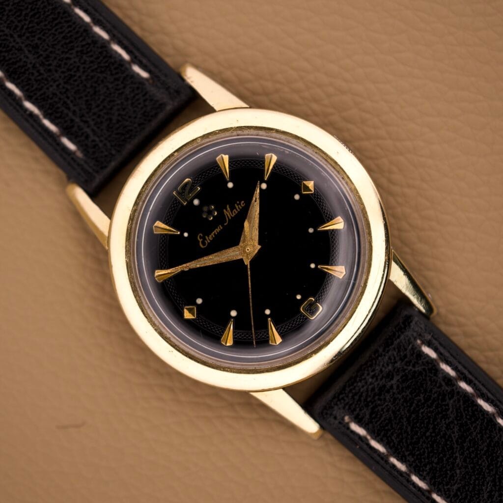 An Eterna Matic Black Dial 14k Gold Capped Case wristwatch with a black leather strap rests on a tan textured surface.