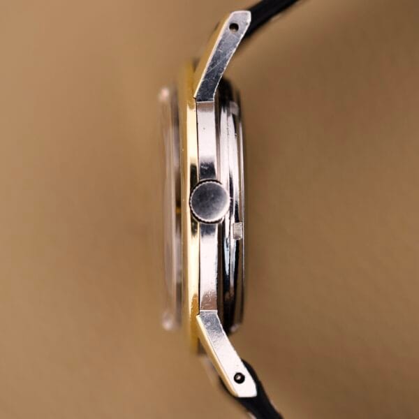Close-up side view of the Eterna Matic Black Dial 14k Gold Capped Case wristwatch, featuring a black dial, black strap, and gold-capped case on a beige background.