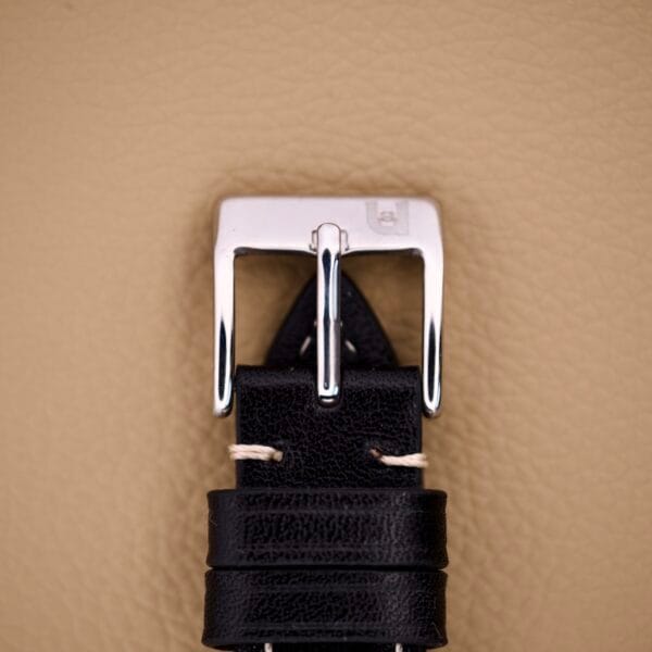 Close-up of a black leather watch strap with a silver buckle on a beige textured background, perfectly matching the Eterna Matic Black Dial 14k Gold Capped Case.