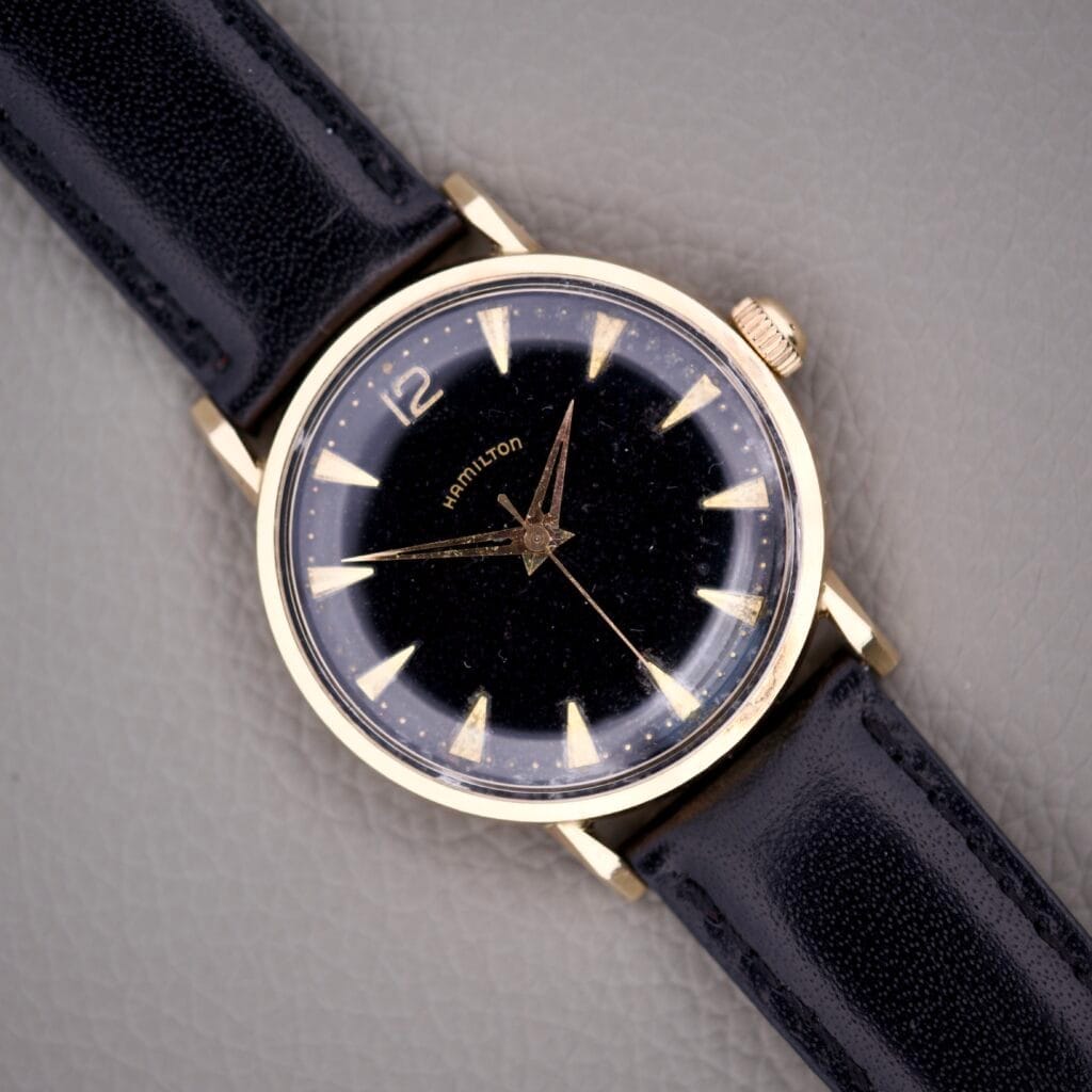 The Hamilton Caliber 671 Manual Wind Black Gilt Dial wristwatch features a gold case, black gilt dial, and black leather strap, displayed on a gray textured surface.