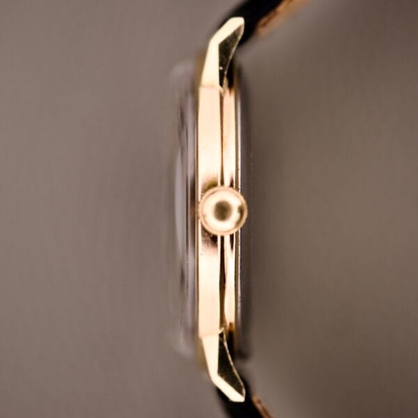 Close-up side view of the Hamilton Caliber 671 Manual Wind with a gold crown, case, and Black Gilt Dial against a neutral background.