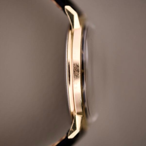 Close-up side view of the Hamilton Caliber 671 Manual Wind with a gold case showing visible scratches, black gilt dial, and a black watch strap.