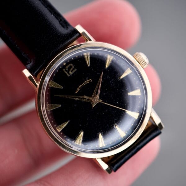 A hand holds the Hamilton Caliber 671 Manual Wind, showcasing its black gilt dial and gold markers.