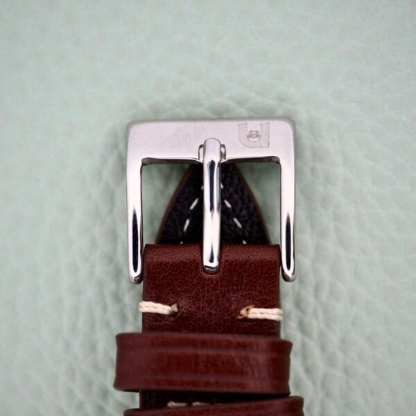 Close-up of a brown leather strap with stainless steel buckle on a light textured background, ideal for pairing with your Benrus 3 Star Automatic #7001 Series Stainless Steel watch.