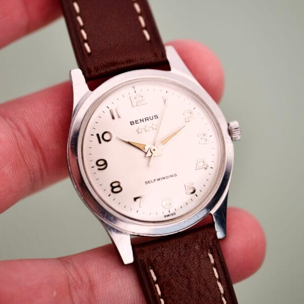 A hand holding the Benrus 3 Star Automatic #7001 Series Stainless Steel watch, featuring a cream dial and brown leather strap.