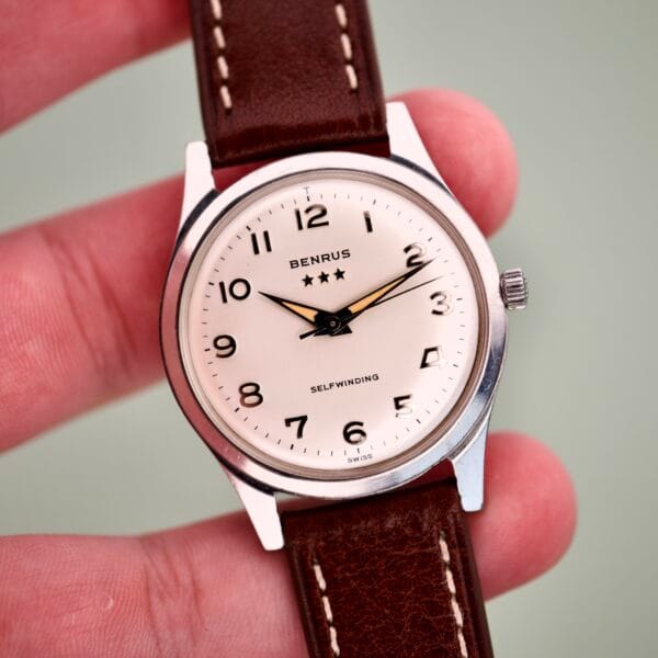 A hand holds the Benrus 3 Star Automatic #7001 Series, featuring a stainless steel case, brown leather strap, and white dial with black numbers.