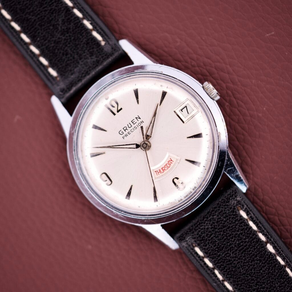 A Gruen Precision Day / Date 522 RSS Sunburst Dial wristwatch with a day-date display and black leather strap rests on a textured brown surface.