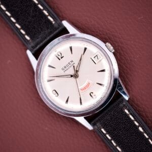 A Gruen Precision Day / Date 522 RSS Sunburst Dial wristwatch with a day-date display and black leather strap rests on a textured brown surface.