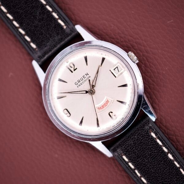 A Gruen Precision Day / Date 522 RSS Sunburst Dial wristwatch with a day-date display and black leather strap rests on a textured brown surface.