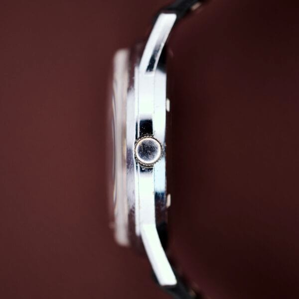 Side view of a silver Gruen Precision Day / Date 522 RSS wristwatch with a sunburst dial and round crown, set against a brown background.