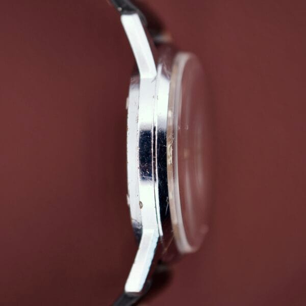 Side view of the Gruen Precision Day / Date 522 RSS Sunburst Dial wristwatch with a silver case, black strap, and sunburst dial, shown against a reddish-brown background.
