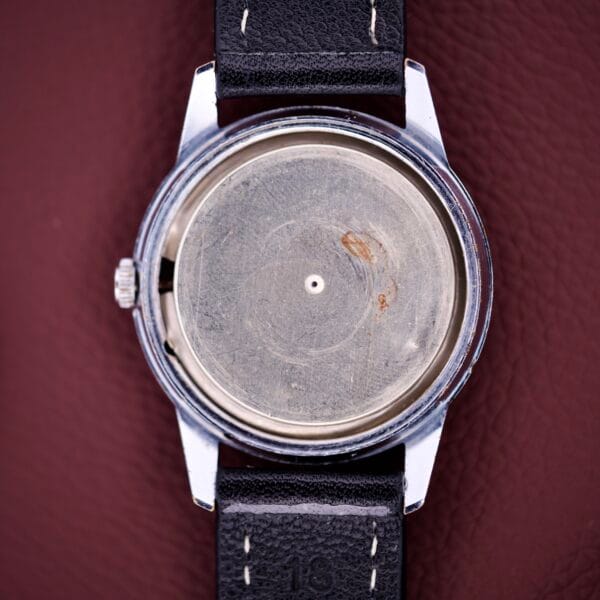A Gruen Precision Day / Date 522 RSS Sunburst Dial wristwatch, missing its face and hands to reveal a blank metal interior, rests on a brown leather surface.
