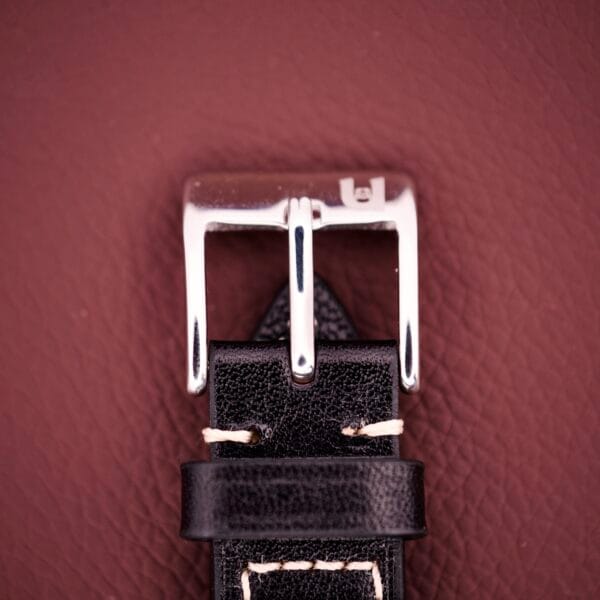 Close-up of a silver buckle on a black leather strap with white stitching, set against a brown background—an ideal match for the classic Gruen Precision Day / Date 522 RSS Sunburst Dial.