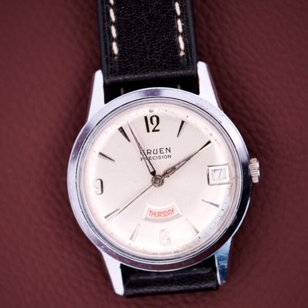 A Gruen Precision Day / Date 522 RSS wristwatch with a sunburst dial and black leather strap, displayed on a brown textured surface.