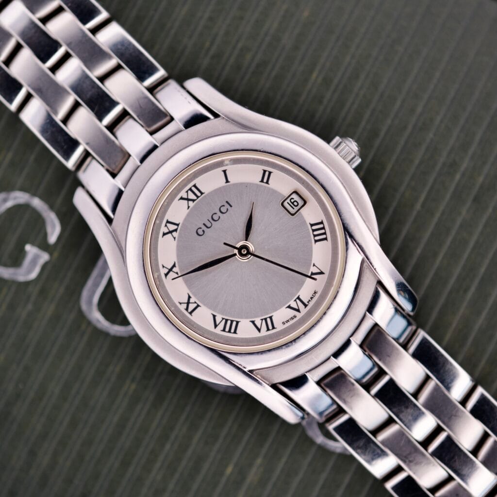 The Gucci 5500L Stainless Steel Quartz wristwatch, featuring Roman numerals and date display, is displayed on a textured dark surface. Includes original box and papers.