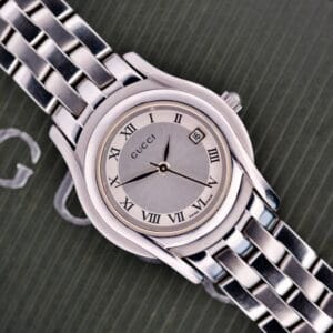 The Gucci 5500L Stainless Steel Quartz wristwatch, featuring Roman numerals and date display, is displayed on a textured dark surface. Includes original box and papers.