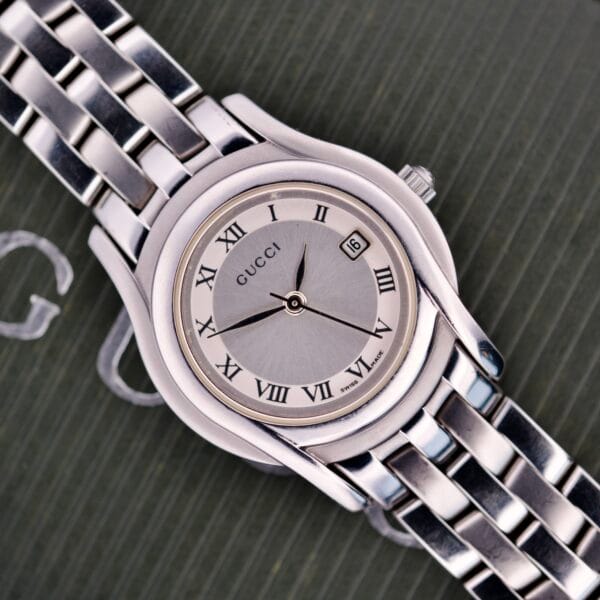 The Gucci 5500L Stainless Steel Quartz wristwatch, featuring Roman numerals and date display, is displayed on a textured dark surface. Includes original box and papers.