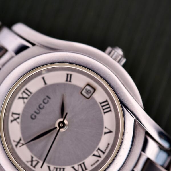 Close-up of a Gucci 5500L Stainless Steel Quartz wristwatch featuring Roman numerals and date display set to the 8th; includes original box and papers.