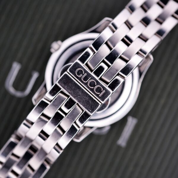 Close-up of a Gucci 5500L Stainless Steel Quartz Box & Papers watch’s bracelet clasp, featuring the brand name elegantly engraved on stainless steel.