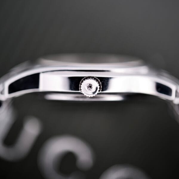 Close-up side view of a Gucci 5500L Stainless Steel Quartz Box & Papers watch crown against a blurred, dark background.