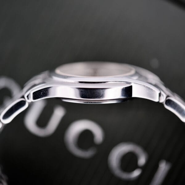 Close-up side view of the Gucci 5500L Stainless Steel Quartz wristwatch, with box and papers, featuring its elegant design and a blurred Gucci logo in the background.