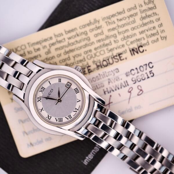 A Gucci 5500L Stainless Steel Quartz Wristwatch with a metal band is displayed with its original box, warranty card, and documentation.