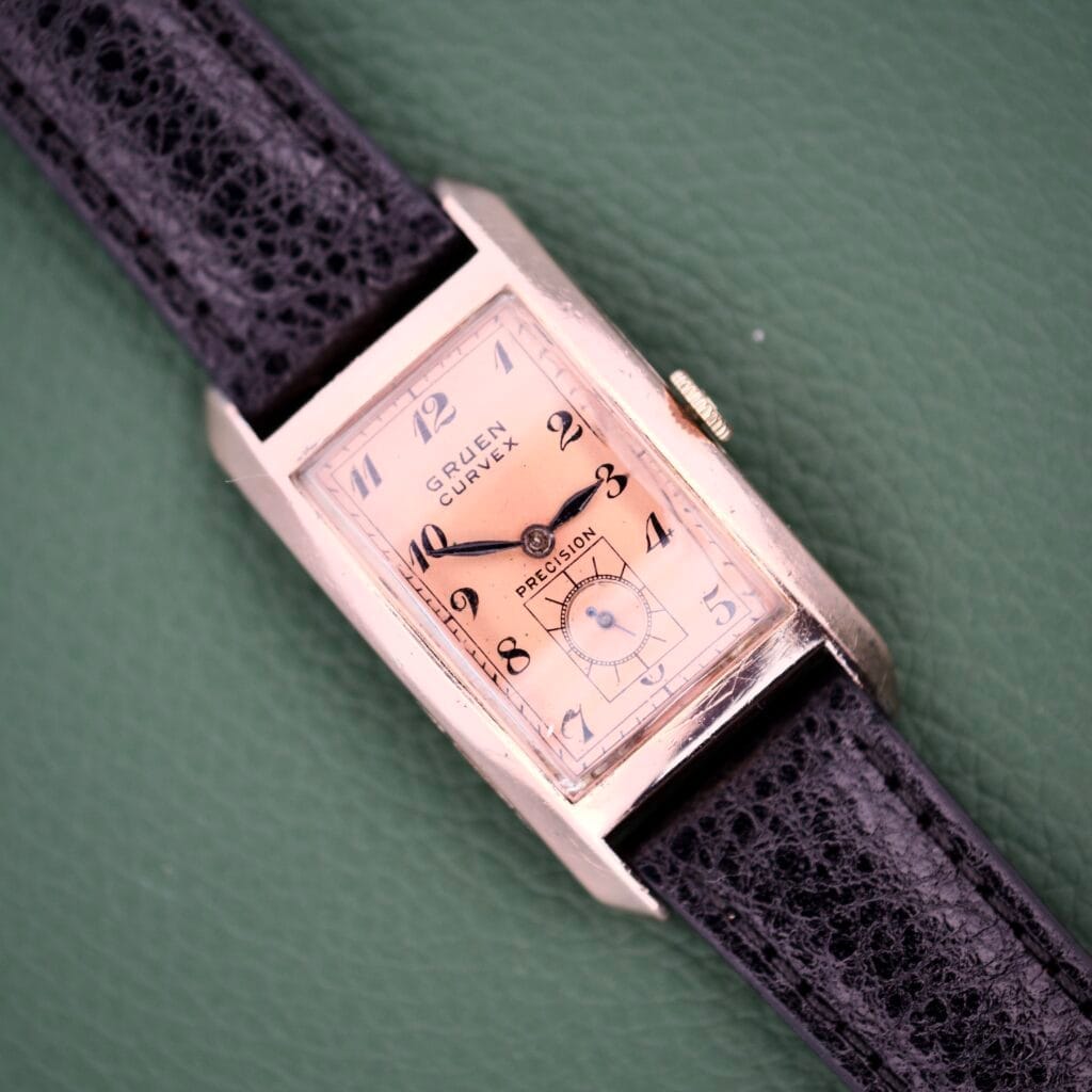 A vintage Gruen Curvex Precision Ref. 440-448 Rose Gold Filled watch with a black leather strap, showcased on a green textured surface.