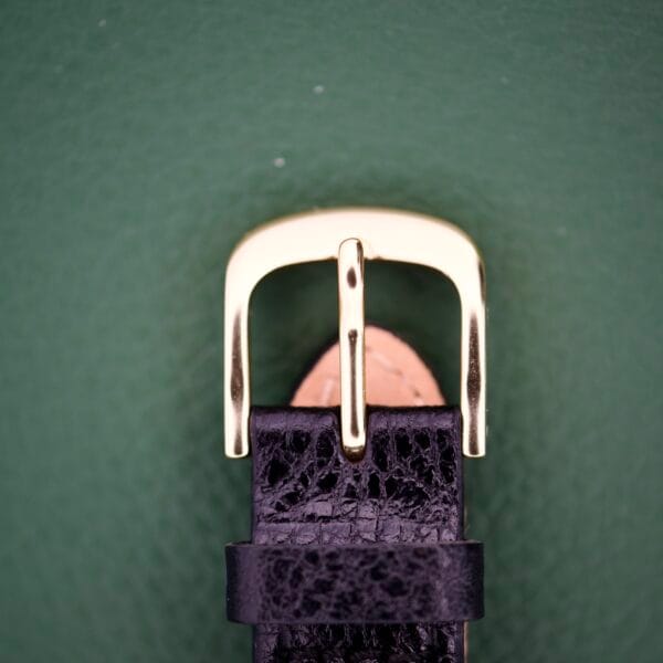Close-up of the Gruen Curvex Precision Ref. 440-448 Rose Gold Filled buckle on a black leather watch strap, set against a green background.