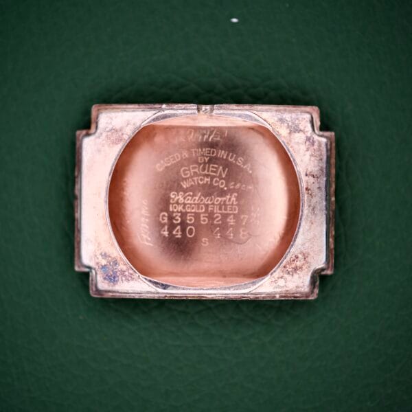 Rectangular vintage Gruen Curvex Precision Ref. 440-448 Rose Gold Filled watch case back with engraved Gruen Watch Co. details, displayed on a green background.